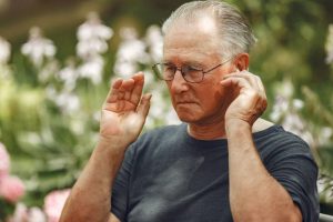 An elderly man outdoors grimacing and touching his ear, illustrating ear discomfort or tinnitus often experienced by seniors.