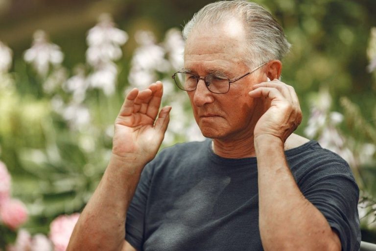 An elderly man outdoors grimacing and touching his ear, illustrating ear discomfort or tinnitus often experienced by seniors.