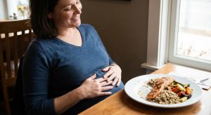 A smiling pregnant woman holding her belly in satisfaction after eating a healthy meal of grilled salmon, quinoa, and roasted vegetables at a cafe table.