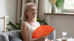A smiling middle-aged woman with blonde hair sits on a grey sofa, holding an open orange hand fan and looking comfortable and relieved, with a glass of ice water on a side table next to her.