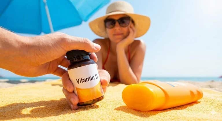 A hand holding a bottle of Vitamin D supplements on a sandy beach towel next to a bottle of sunscreen, with a woman sunbathing under an umbrella in the background, illustrating the question of supplementing during summer.
