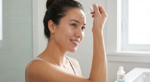 A smiling woman with her hair pulled back uses a glass dropper to apply a clear liquid from a bottle labeled "HYDRATE + SAFE SERUM" onto her forehead in a bright bathroom setting.