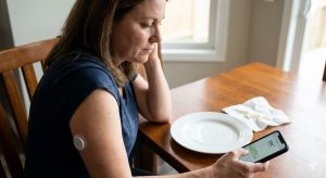 A woman sits at a dining table with an empty plate, looking at continuous glucose monitor (CGM) readings on her smartphone, with a sensor visible on her arm.