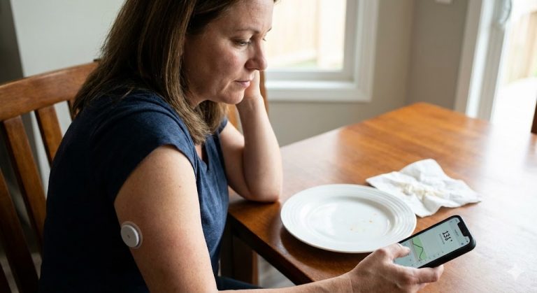 A woman sits at a dining table with an empty plate, looking at continuous glucose monitor (CGM) readings on her smartphone, with a sensor visible on her arm.