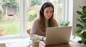 A young woman with a calm, focused expression, wearing headphones and a knitted sweater, works on a silver laptop at a wooden desk by a large window, representing the "flow state."