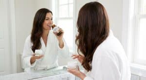 A woman in a bathrobe looking in a bathroom mirror, simultaneously taking an oral supplement from an amber bottle and applying a topical cream from a white jar to her hair, illustrating the combined internal and external approach to hair health.