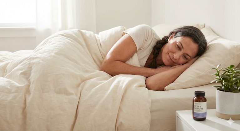 A woman sleeps peacefully in a sunlit bed with a "Rest & Restore" supplement bottle on the nightstand.