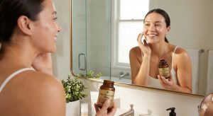 A woman with clear, glowing skin smiles at her reflection in a bathroom mirror while holding a bottle of Zinc Picolinate 50mg supplement, suggesting its benefits for acne and overall skin health.