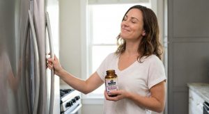 A relieved woman smiles while holding a bottle of Daily Woman's Probiotic taken from a refrigerator in a kitchen.