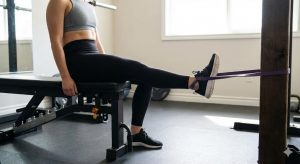 A woman seated on a gym bench performs a terminal knee extension exercise using a resistance band anchored to a post.